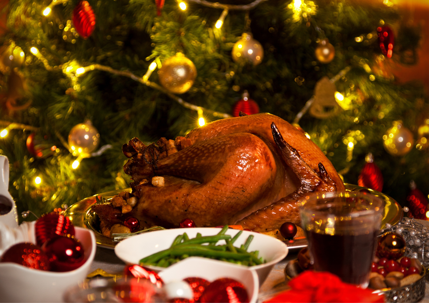 A beautifully roasted turkey sits as the centerpiece of a festive holiday table, surrounded by green beans, cranberries, and a glass of red wine. In the background, a Christmas tree glows warmly with golden lights and ornaments in shades of red and gold, creating a cozy and celebratory atmosphere. Bright decorations and a cheerful ambience make the setting perfect for a holiday feast.