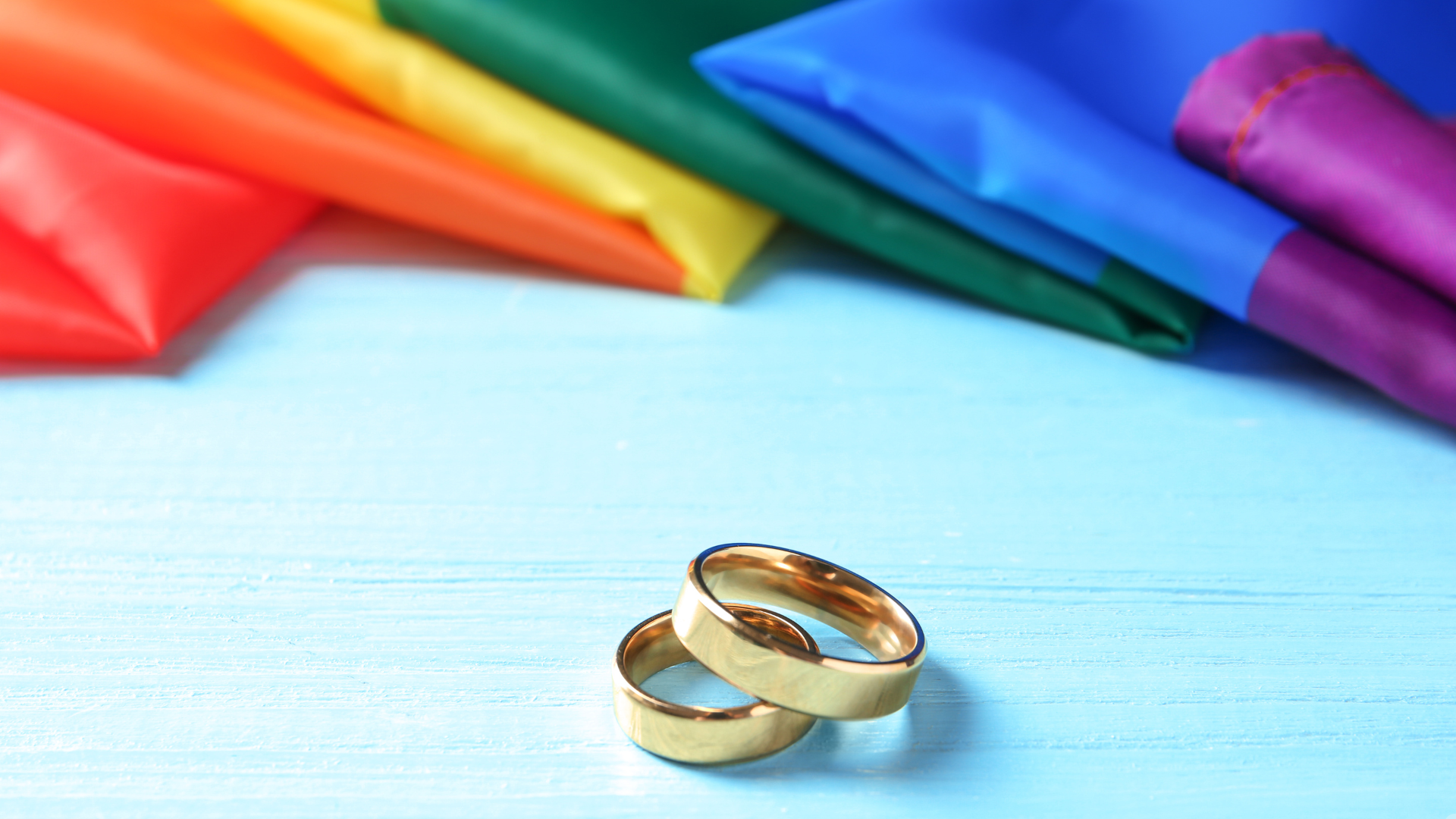 Two gold wedding rings rest on a light blue wooden surface, with a vibrant rainbow-colored fabric draped in the background, symbolizing LGBTQ+ pride and love